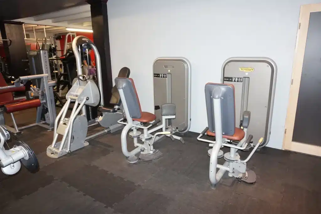 Salle de sport avec plusieurs appareils de musculation gris et marron. Au premier plan, deux machines Technogym Adduction.