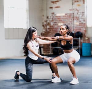Coach sportive brune guide une femme réalisant un squat, bras tendus, pour corriger la posture en salle de gym.