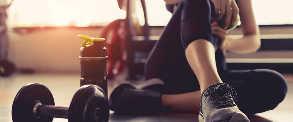 Femme en tenue de sport assise au sol dans une salle de gym, tenant une pomme verte. Un haltère et une bouteille shaker noirs sont à côté.
